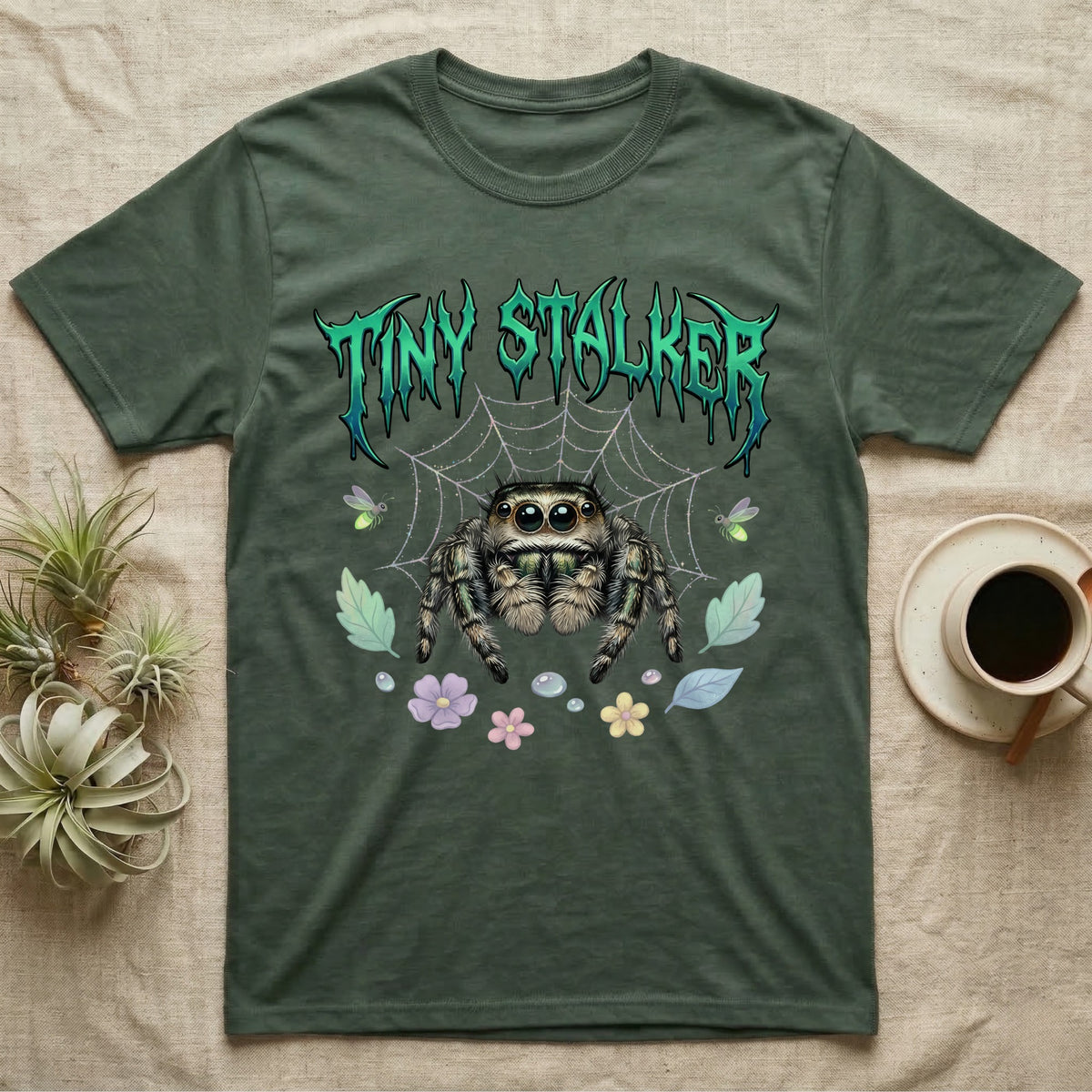Regal Jumping Spider Feral T-Shirt in Military Green — Fuzzy jumping spider with huge eyes in a pastel garden under a spiky iridescent-teal metal wordmark reading TINY STALKER.
