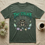 Regal Jumping Spider Feral T-Shirt in Military Green — Fuzzy jumping spider with huge eyes in a pastel garden under a spiky iridescent-teal metal wordmark reading TINY STALKER.