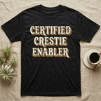 Certified Crestie Enabler T-Shirt in Black — Bold retro drop-shadow wordmark reading "Certified Crestie Enabler" in cream and ochre on a grey background.