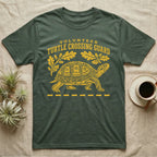 Box Turtle Crossing T-Shirt in Military Green — Yellow folk-print eastern box turtle walking across a dashed road line with oak leaves, text Volunteer Turtle Crossing Guard