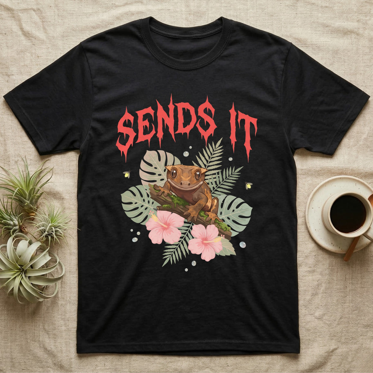 Crested Gecko Feral T-Shirt in Black — Crested gecko on a mossy branch in a pastel jungle scene under a spiky coral metal wordmark reading SENDS IT.