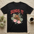 Crested Gecko Feral T-Shirt in Black — Crested gecko on a mossy branch in a pastel jungle scene under a spiky coral metal wordmark reading SENDS IT.