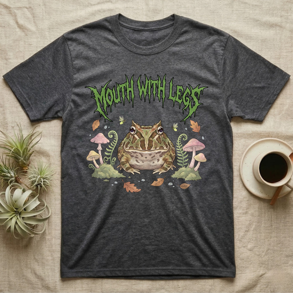 Pacman Frog Feral T-Shirt in Dark Heather — Round Pacman frog in a pastel forest-floor scene under a spiky leaf-green metal wordmark reading MOUTH WITH LEGS.