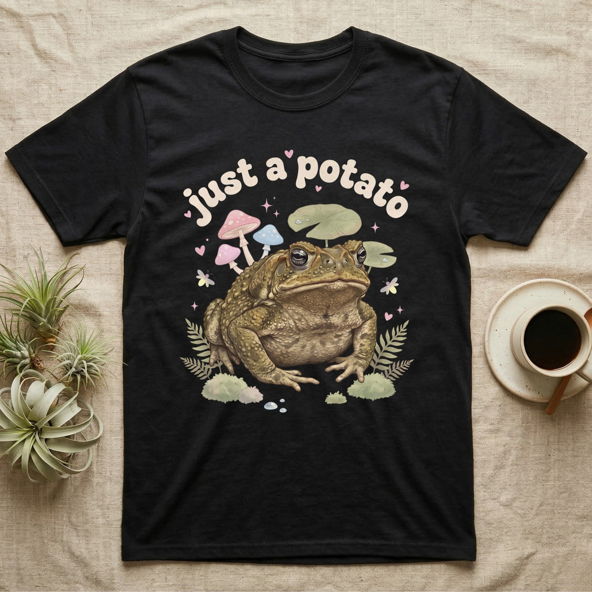 Cane Toad Baby T-Shirt in Black — Warty cane toad among pastel toadstools and lily pads under puffy cream bubble text "just a potato".
