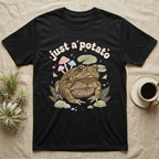 Cane Toad Baby T-Shirt in Black — Warty cane toad among pastel toadstools and lily pads under puffy cream bubble text "just a potato".