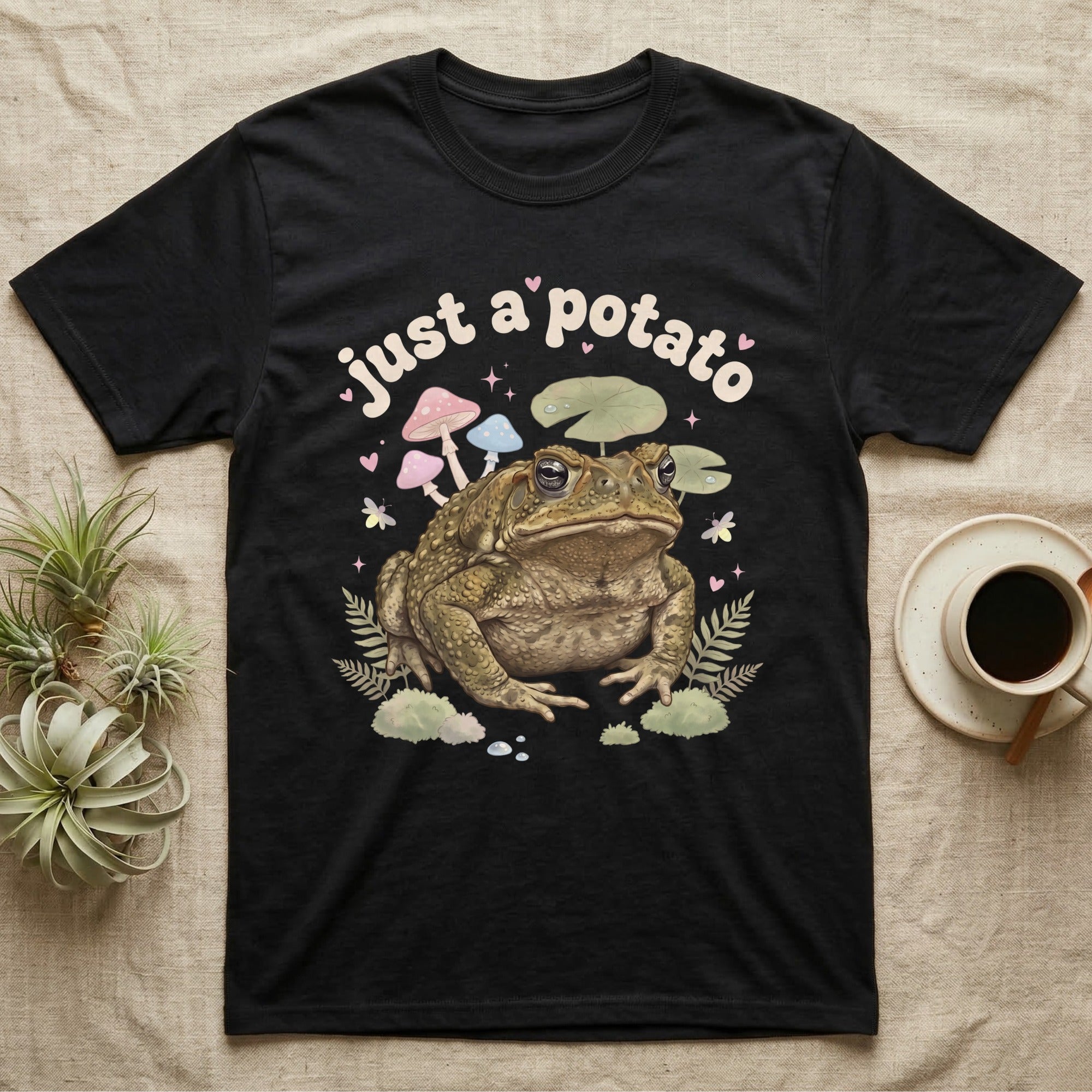 Cane Toad Baby T-Shirt in Black — Warty cane toad among pastel toadstools and lily pads under puffy cream bubble text "just a potato".