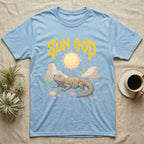 Moroccan Uromastyx Feral T-Shirt in Light Blue — Moroccan uromastyx basking under a big sun with a jagged solar-gold metal "SUN GOD" wordmark and pastel desert scene.