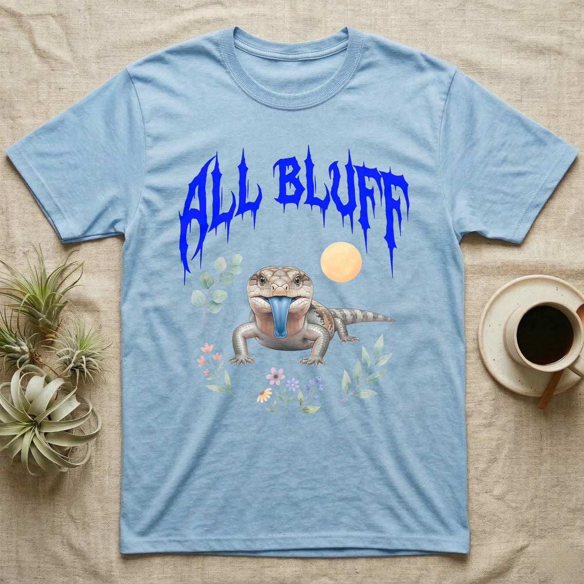 Northern Blue-Tongue Skink Feral T-Shirt in Light Blue — Blue-tongue skink with its blue tongue out under a jagged cobalt-blue metal "ALL BLUFF" wordmark and pastel leaves.
