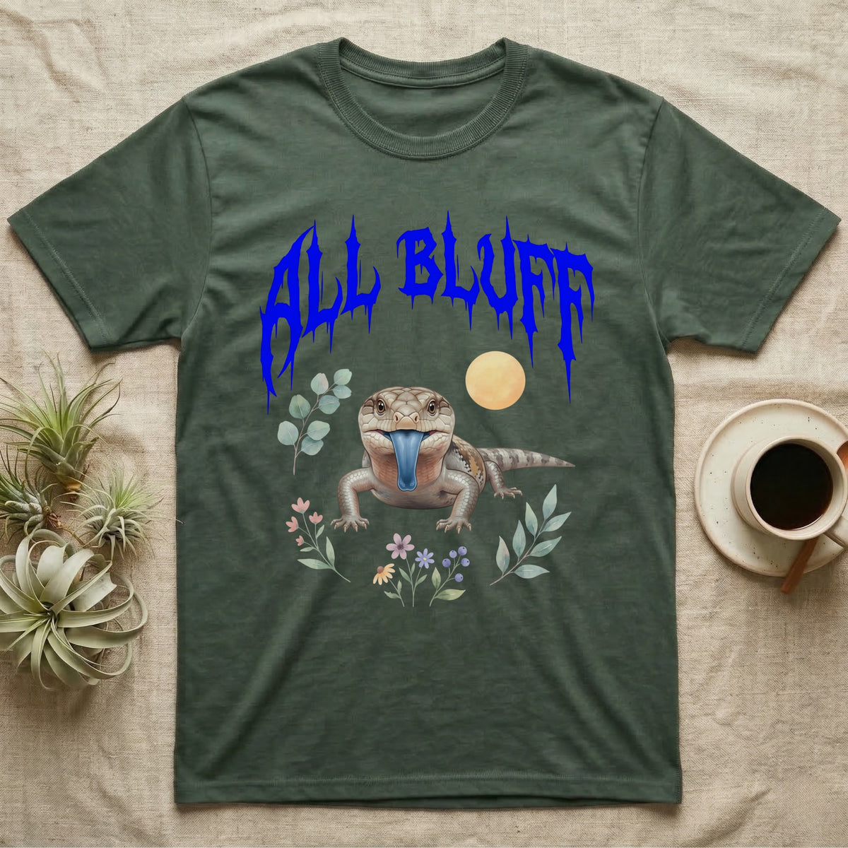 Northern Blue-Tongue Skink Feral T-Shirt in Military Green — Blue-tongue skink with its blue tongue out under a jagged cobalt-blue metal "ALL BLUFF" wordmark and pastel leaves.