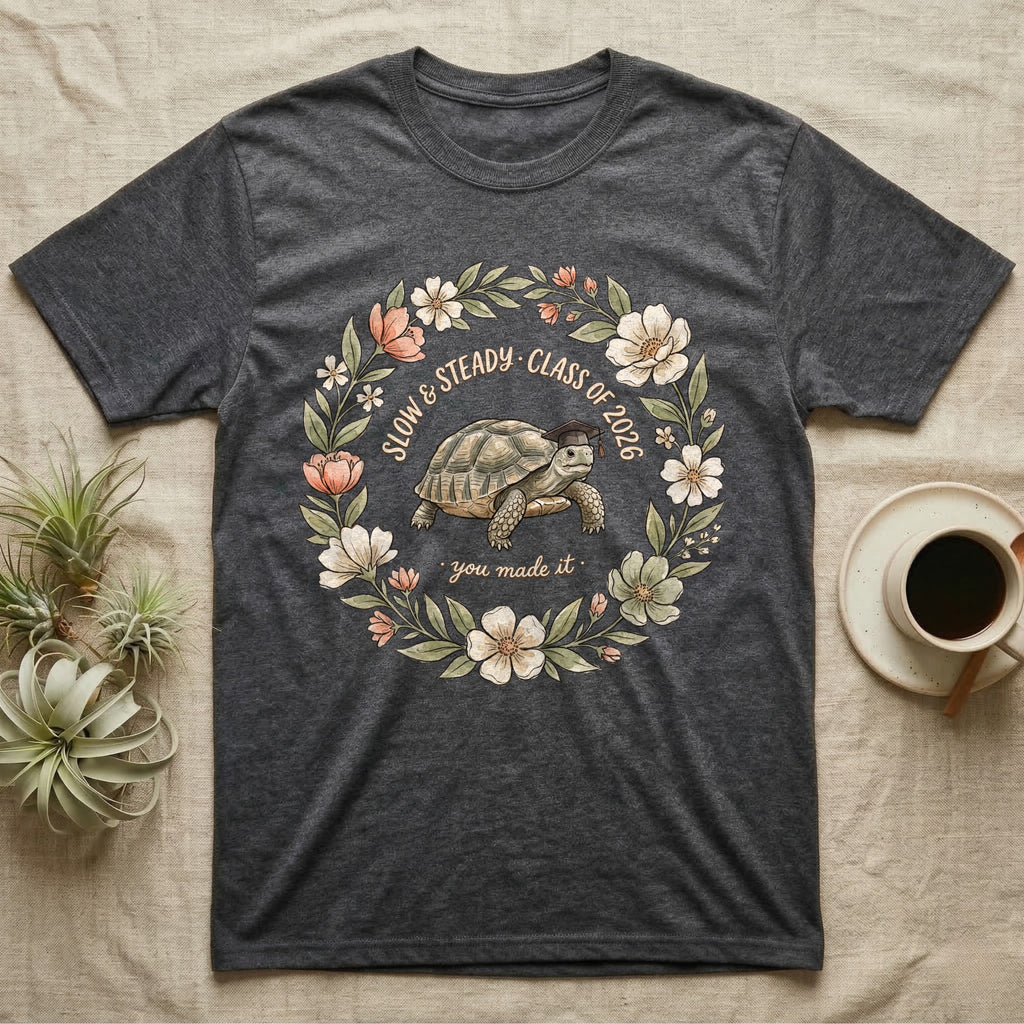 Slow And Steady Graduation T-Shirt in Dark Heather — Watercolour illustration of a tortoise in a graduation cap inside a floral wreath with "Slow & Steady, Class of 2026."