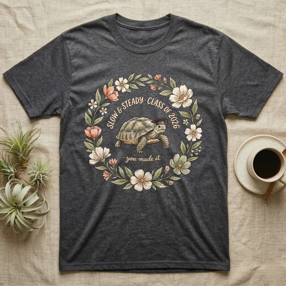 Slow And Steady Graduation T-Shirt in Dark Heather — Watercolour illustration of a tortoise in a graduation cap inside a floral wreath with "Slow & Steady, Class of 2026."