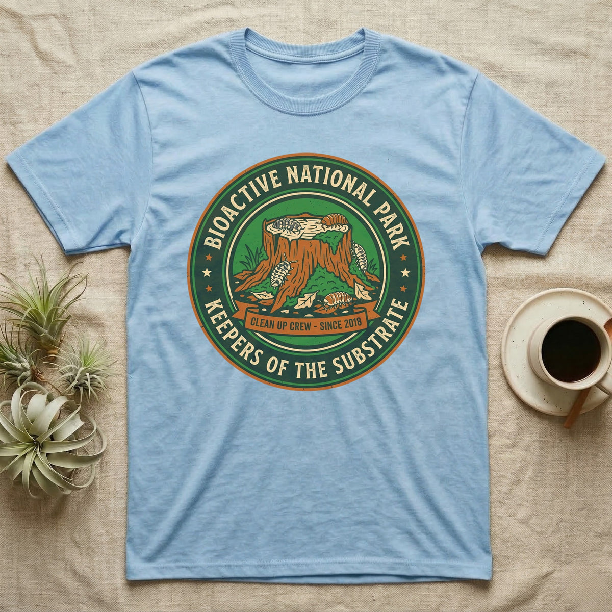 Isopod Keepers of the Substrate T-Shirt in Light Blue — Weathered green park badge with isopods working a mossy stump and leaf litter, cream arc lettering.