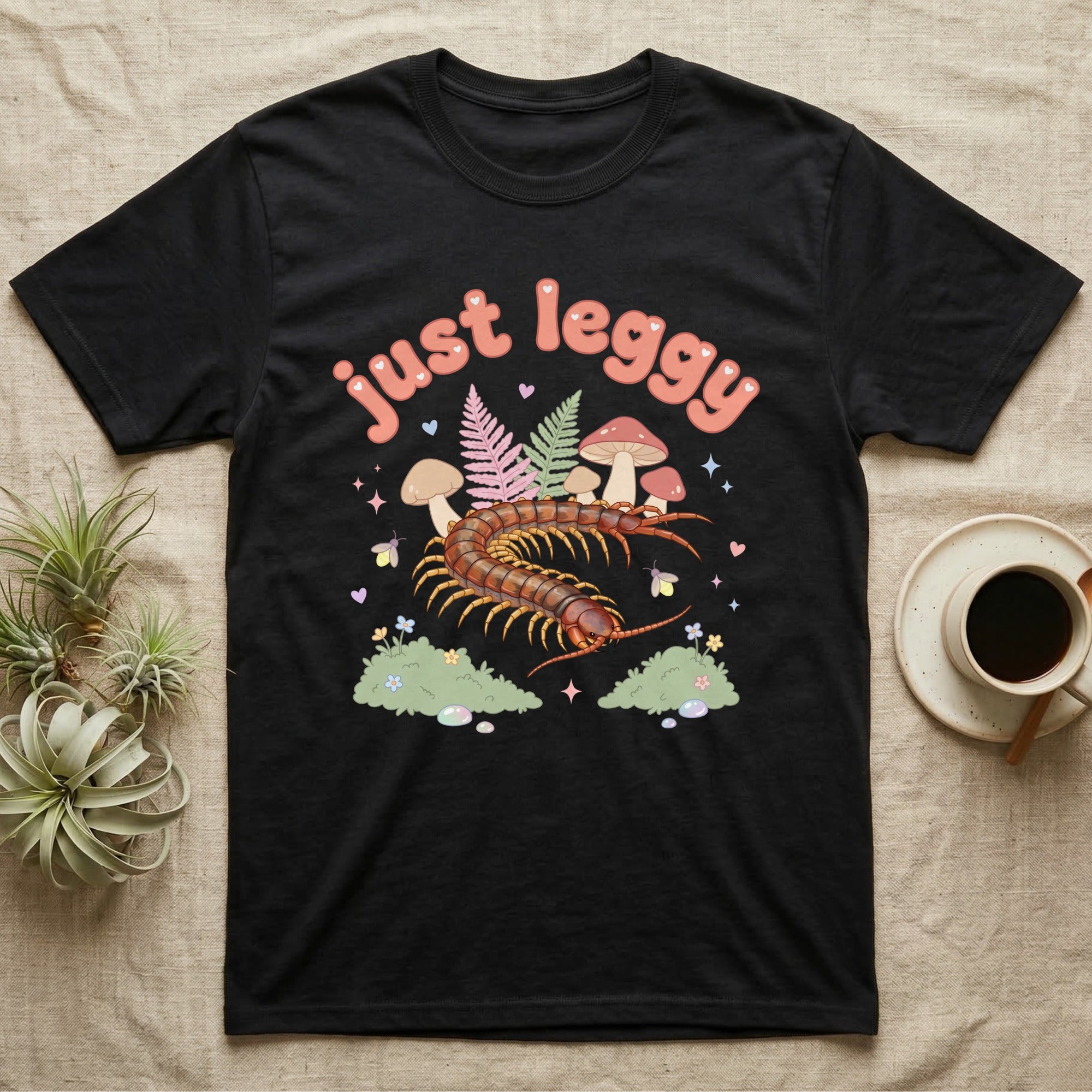 Vietnamese Centipede Baby T-Shirt in Black — Many-legged Vietnamese centipede curving among pastel ferns and toadstools under puffy coral text "just leggy".