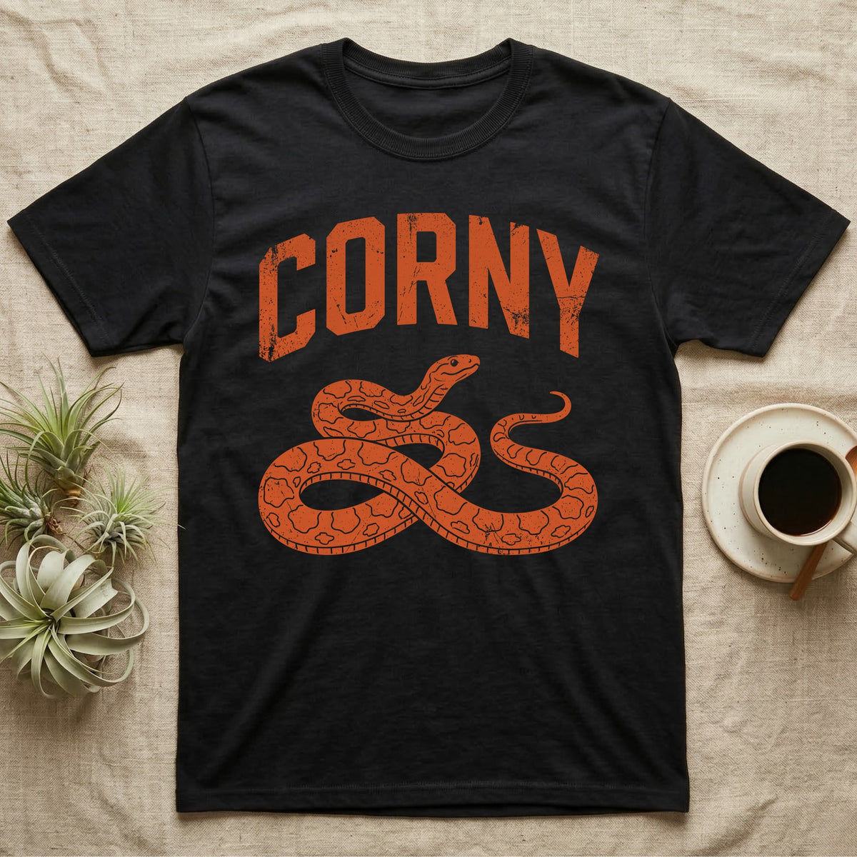 Corn Snake Corny T-Shirt in Black — Worn orange one-ink print of a corn snake lying in loose coils, head raised, under the arched word Corny