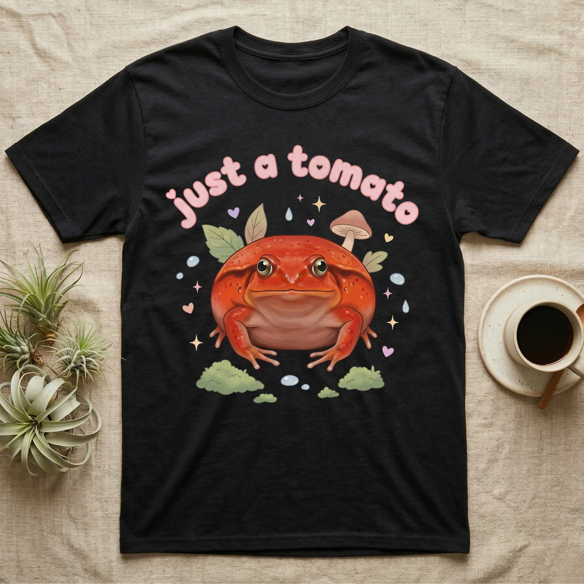 Tomato Frog Softie T-Shirt in Black — Round red-orange tomato frog facing forward under a puffy pastel "just a tomato" bubble wordmark with tiny hearts.
