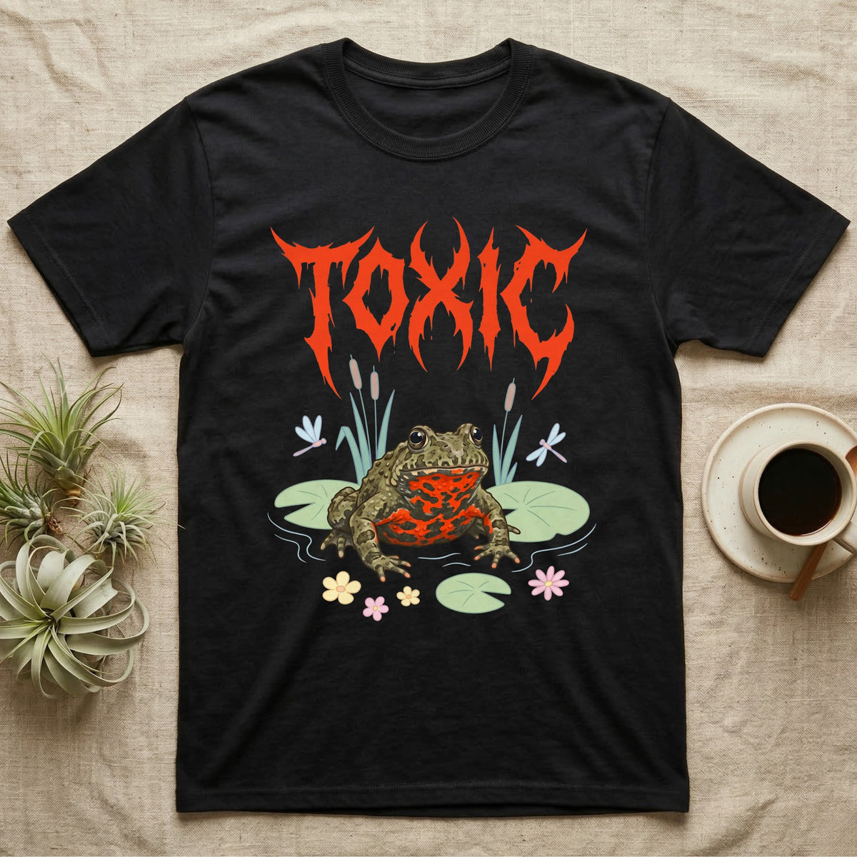 Oriental Fire-Bellied Toad Feral T-Shirt in Black — Pastel fire-bellied toad with red-orange belly among lily pads and dragonflies, jagged metal wordmark "TOXIC" above.