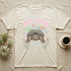 Common Snapping Turtle Baby T-Shirt in Natural — Snapping turtle with hooked beak and spiky shell among pastel lily pads under puffy pink bubble text "just a baby".