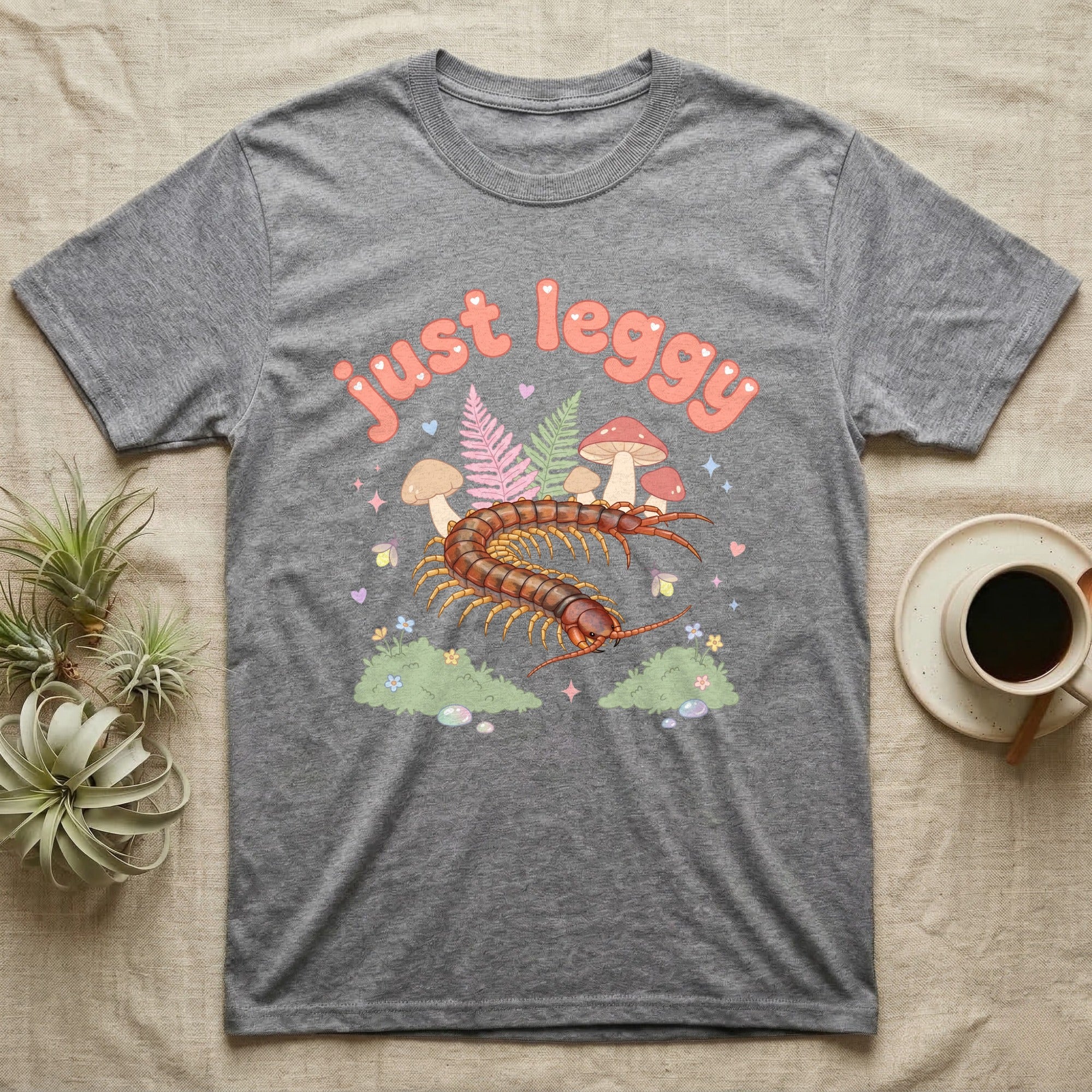 Vietnamese Centipede Baby T-Shirt in Sport Grey — Many-legged Vietnamese centipede curving among pastel ferns and toadstools under puffy coral text "just leggy".