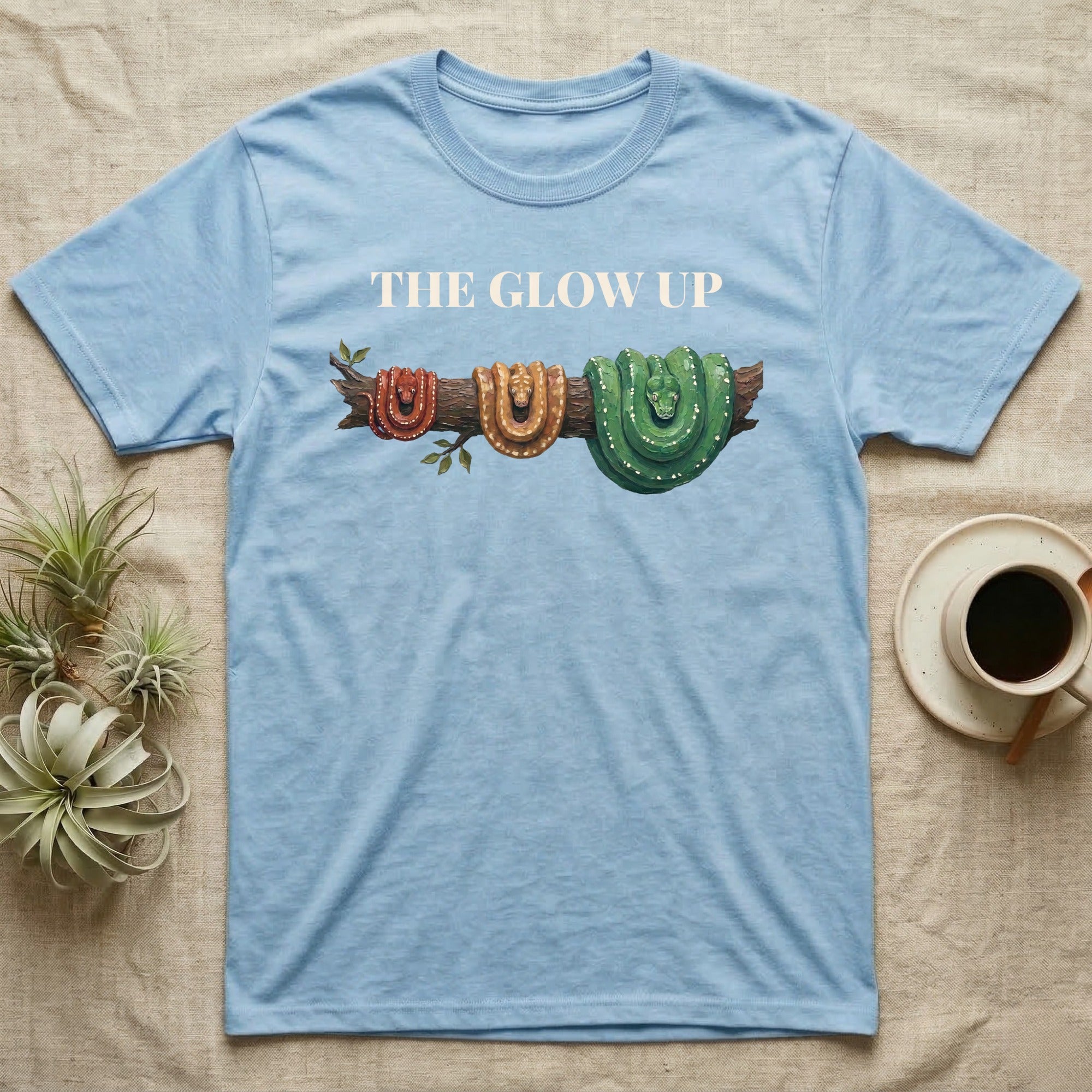 Green Tree Python Glow-Up T-Shirt in Light Blue — Impasto illustration of a green tree python at three life stages on one branch: red hatchling, orange-brown subadult, green adult.