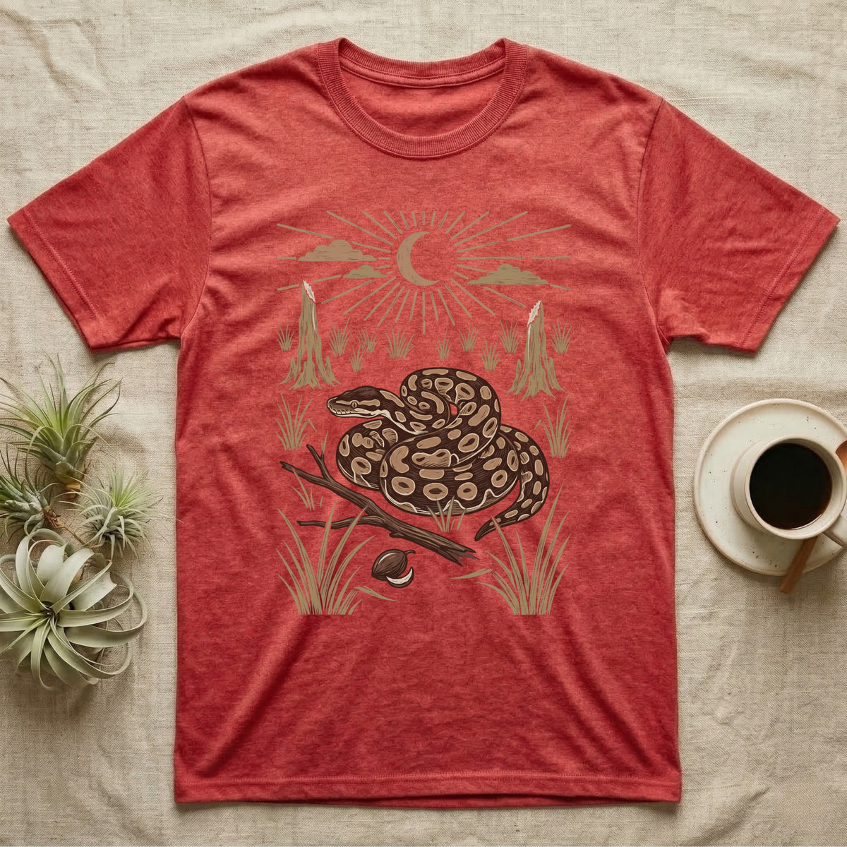 Ball Python Home Range T-Shirt in Heather Red — Woodcut-style illustration of a ball python coiled on dry savanna ground with grass tufts and termite mounds behind, no on-shirt text.