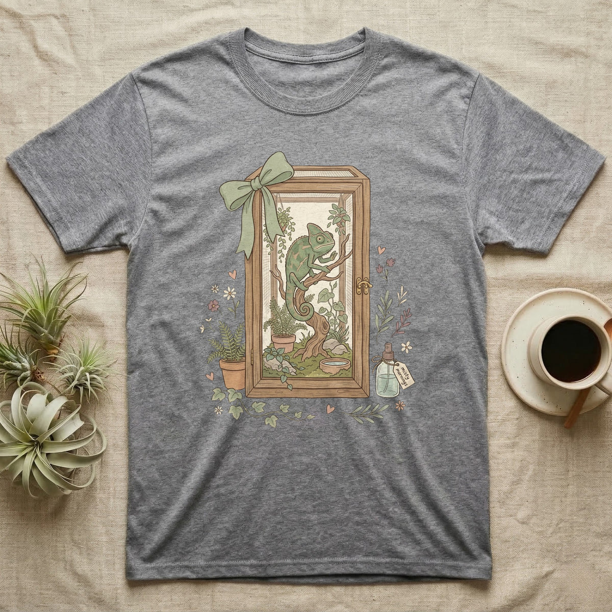 Chameleon in terrarium with plants, framed by a bow, on a grey t-shirt.