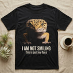 Photographic mugshot of a leopard gecko braced low and staring into the lens above the hook I Am Not Smiling, this is just my face. — shown on a Black tee.