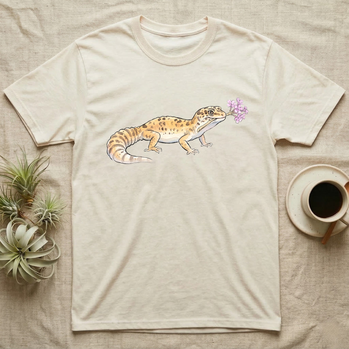 Leopard Gecko Blossom T-Shirt in Natural — Watercolor illustration of a sandy-gold spotted leopard gecko holding a sand verbena blossom sprig, on a dark grey t-shirt.