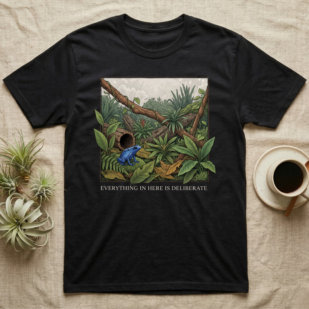 Dart Frog Bioactive Vivarium T-Shirt in Black — Illustrated panel of a cobalt dart frog in a layered planted vivarium, leaves crossing the inked border.