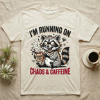 Cream t-shirt with raccoon holding "Chaos Fuel" mug, text "I'M RUNNING ON CHAOS & CAFFEINE".