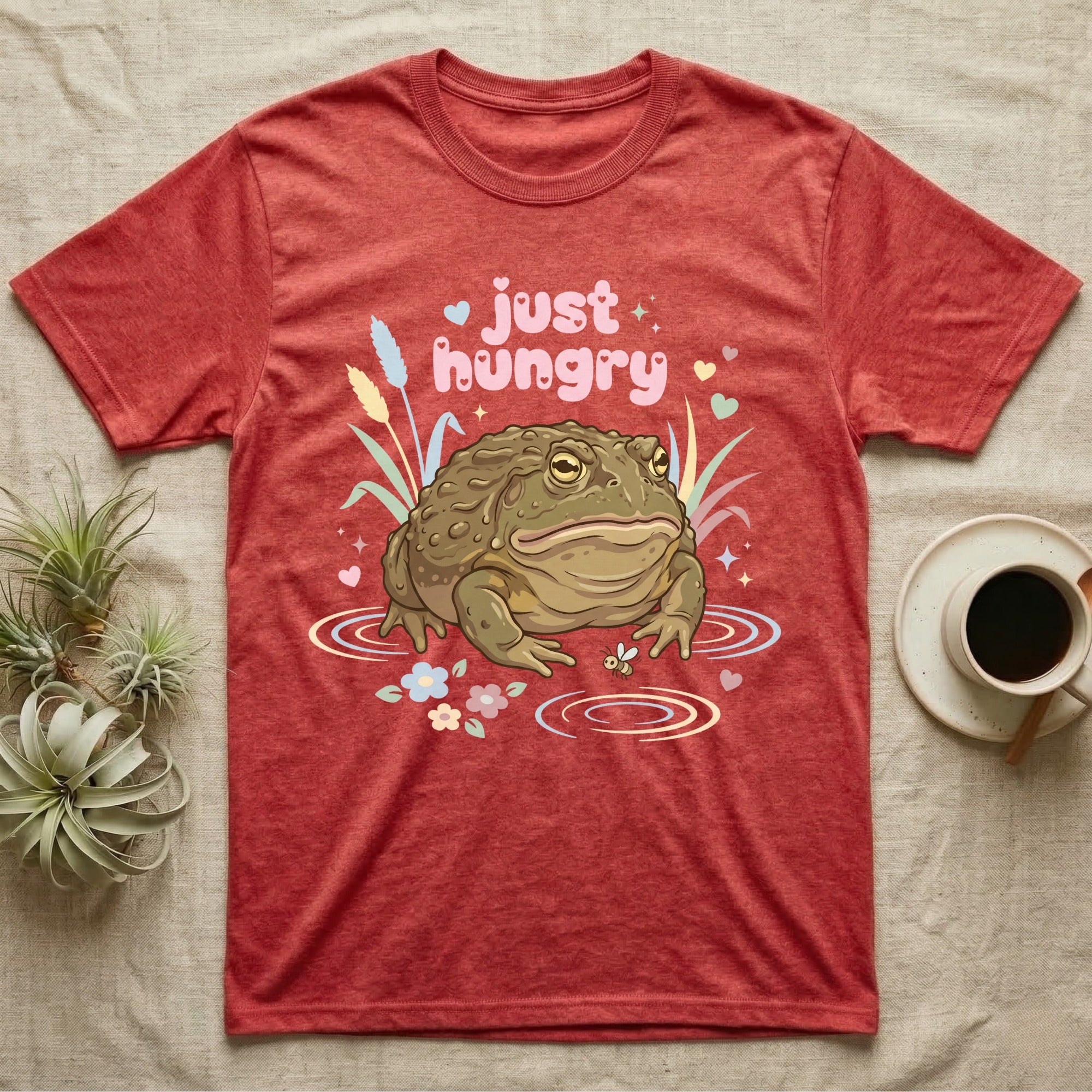 African Bullfrog Baby T-Shirt in Heather Red — Pastel African bullfrog eyeing a tiny bug among reeds and flowers, puffy pink bubble wordmark "just hungry" above.