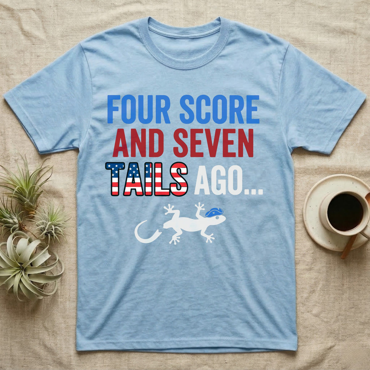 Gecko Four Score T-Shirt in Light Blue — Block text Four Score and Seven Tails Ago with a flag-filled word above a white gecko silhouette shedding its tail.