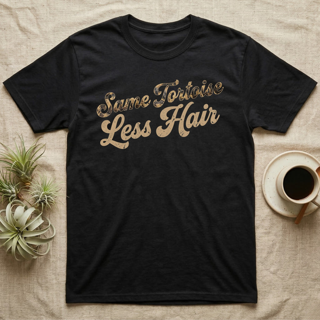 Tortoise Owner Retro T-Shirt in Black — Worn 1970s brush script reading Same Tortoise in tortoise-shell texture over Less Hair in warm sand, climbing a diagonal on charcoal grey