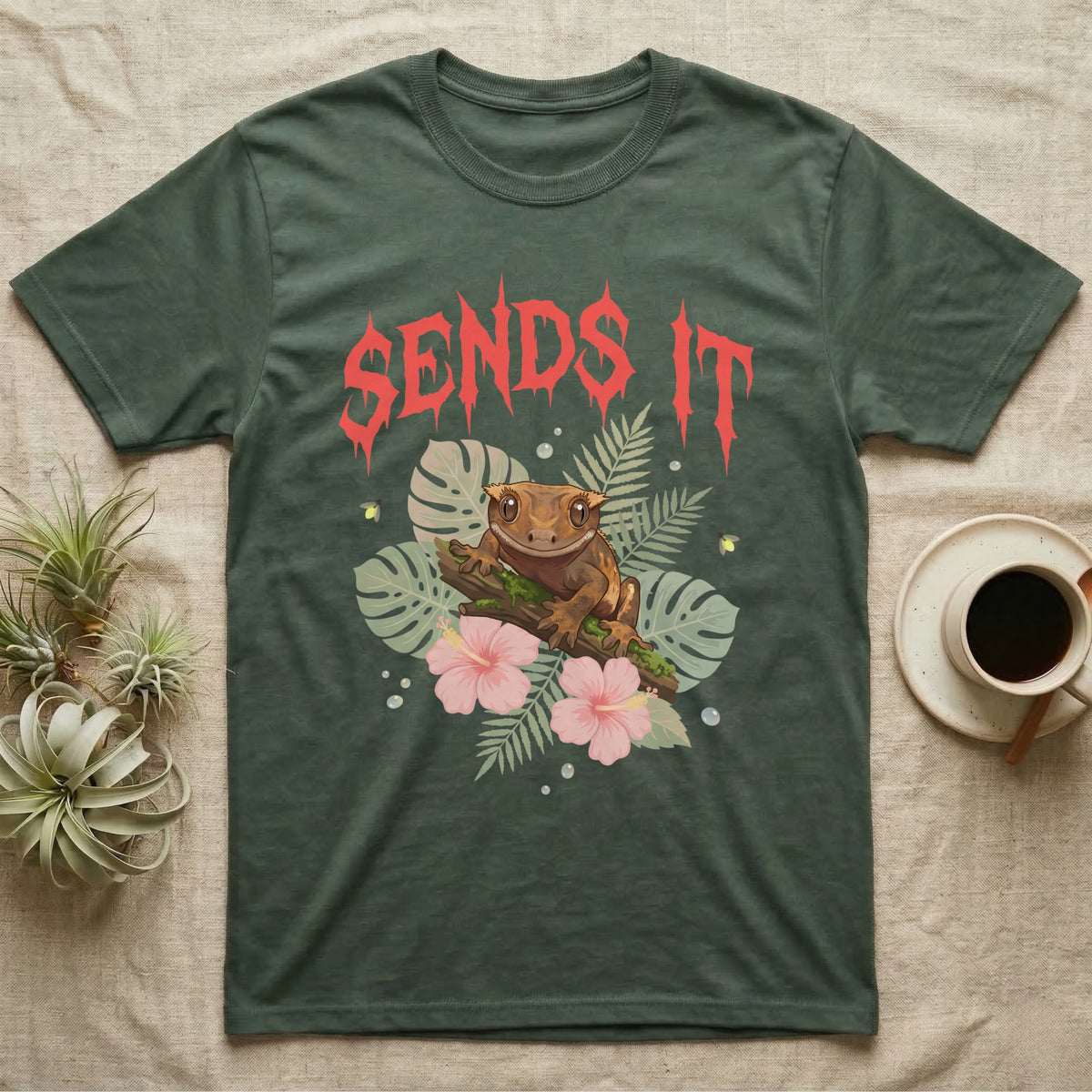 Crested Gecko Feral T-Shirt in Military Green — Crested gecko on a mossy branch in a pastel jungle scene under a spiky coral metal wordmark reading SENDS IT.