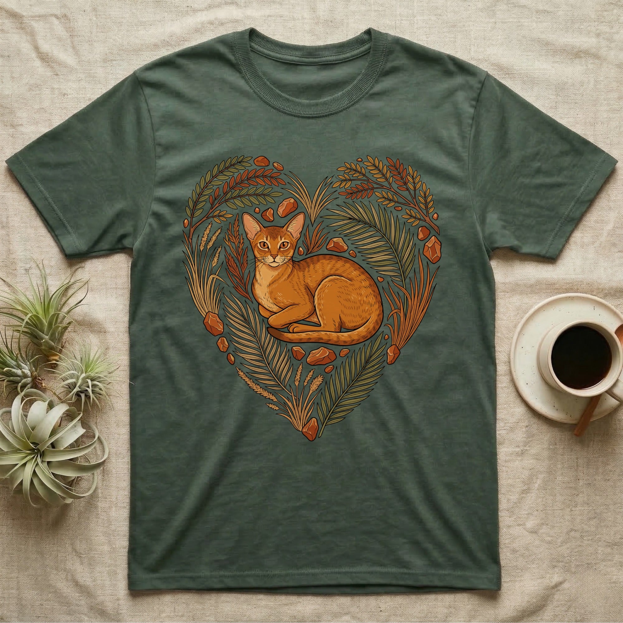 Green t-shirt featuring a stylized cat resting in a heart-shaped arrangement of leaves and stones.