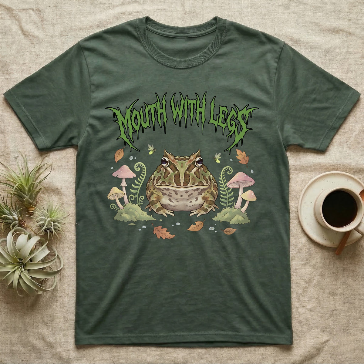 Pacman Frog Feral T-Shirt in Military Green — Round Pacman frog in a pastel forest-floor scene under a spiky leaf-green metal wordmark reading MOUTH WITH LEGS.