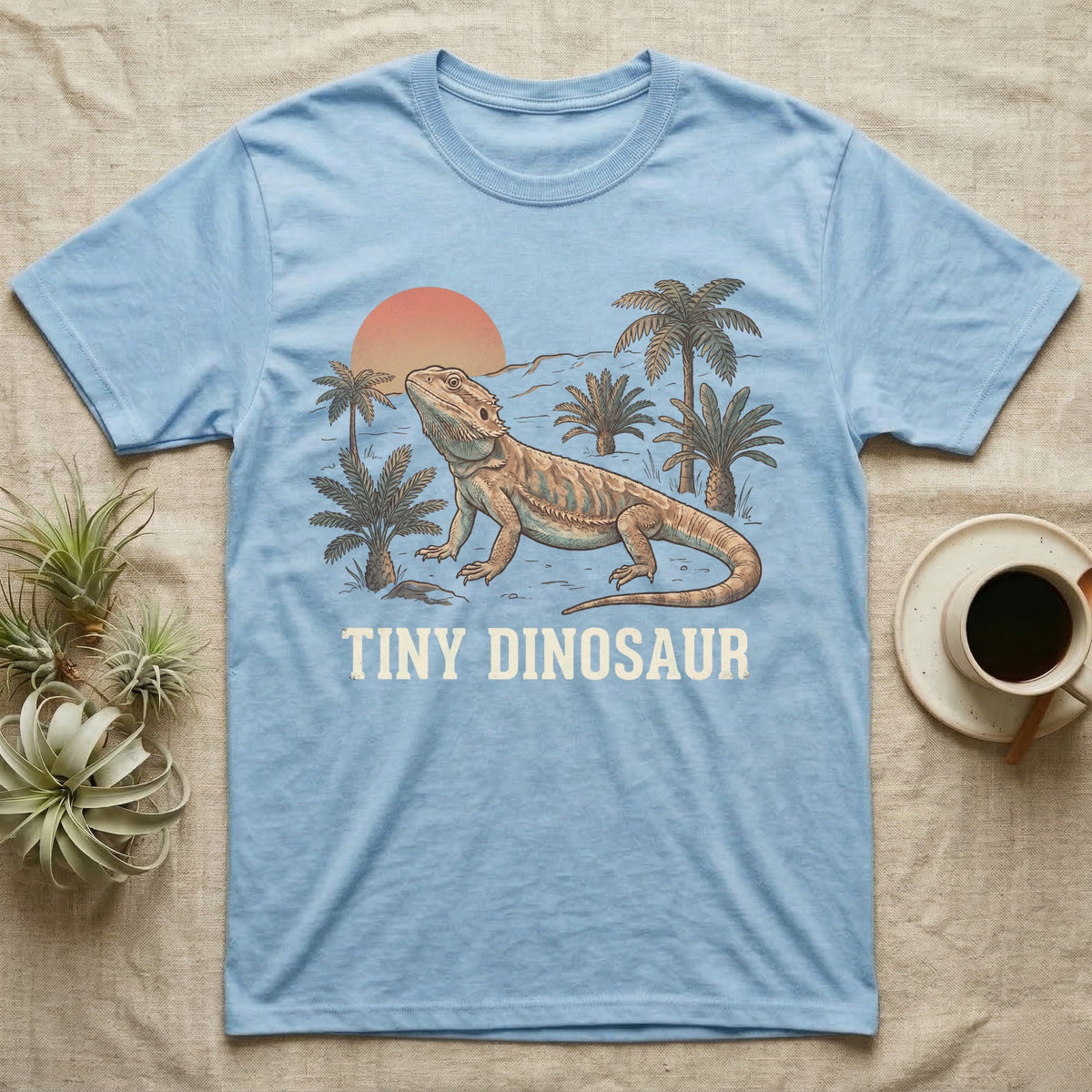 Tiny Dinosaur Bearded Dragon T-Shirt in Light Blue — Retro natural-history illustration of a bearded dragon in profile in a prehistoric landscape with a distressed "Tiny Dinosaur" caption.