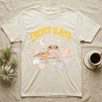 Leopard Gecko Feral T-Shirt in Natural — Smiling leopard gecko in a pastel desert-night scene under a spiky golden metal wordmark reading CRICKET SLAYER.