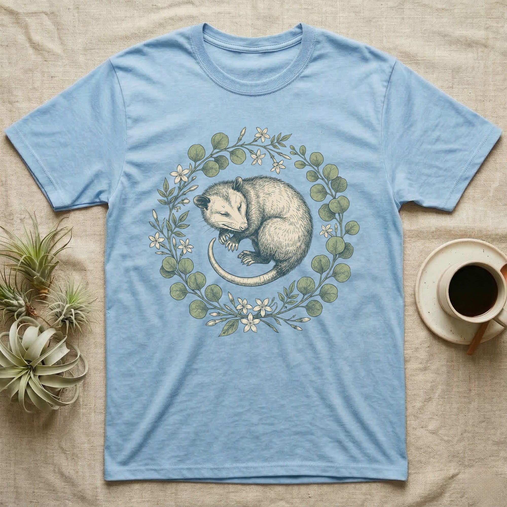 Light blue t-shirt featuring a sleeping opossum surrounded by a floral and leafy wreath.