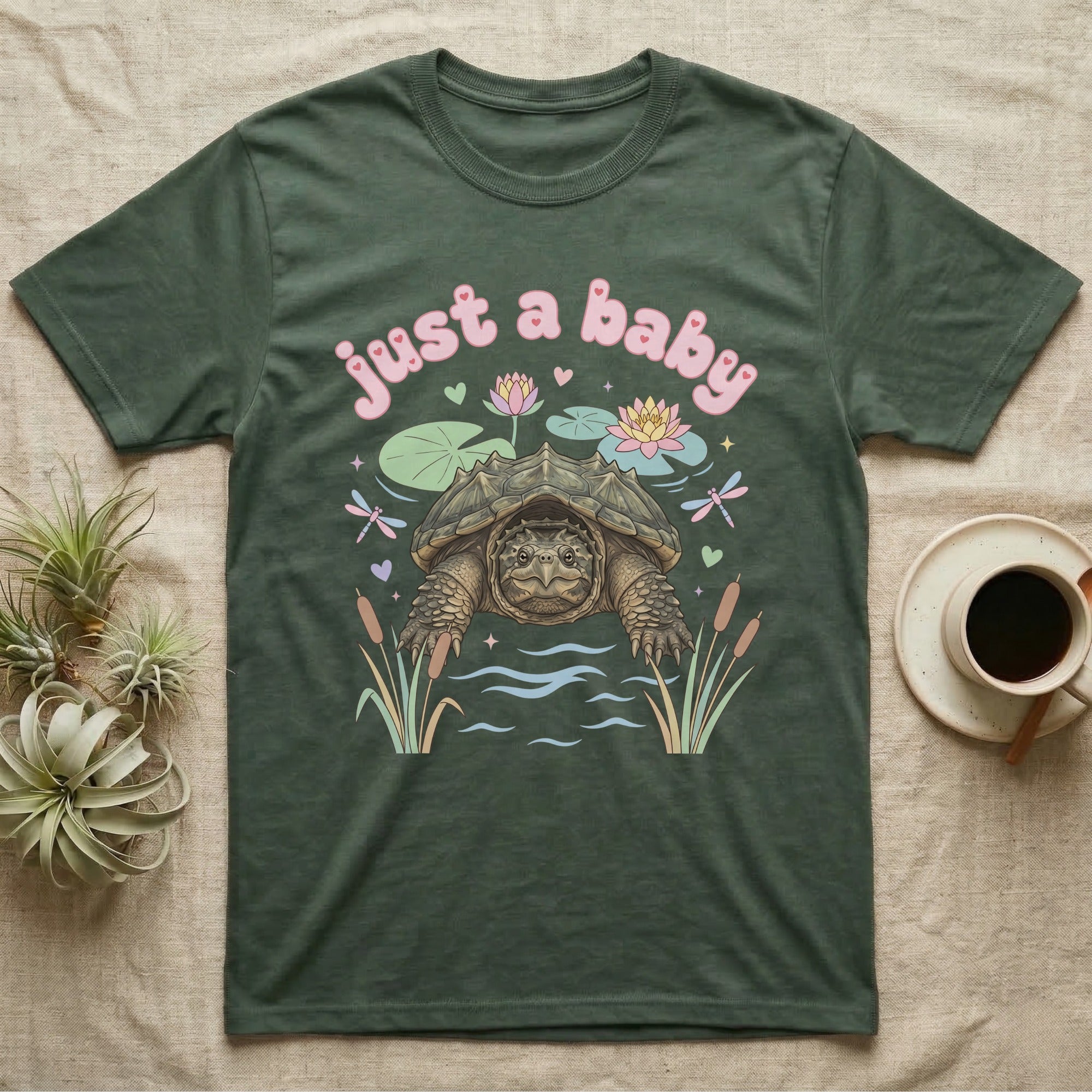 Common Snapping Turtle Baby T-Shirt in Military Green — Snapping turtle with hooked beak and spiky shell among pastel lily pads under puffy pink bubble text "just a baby".
