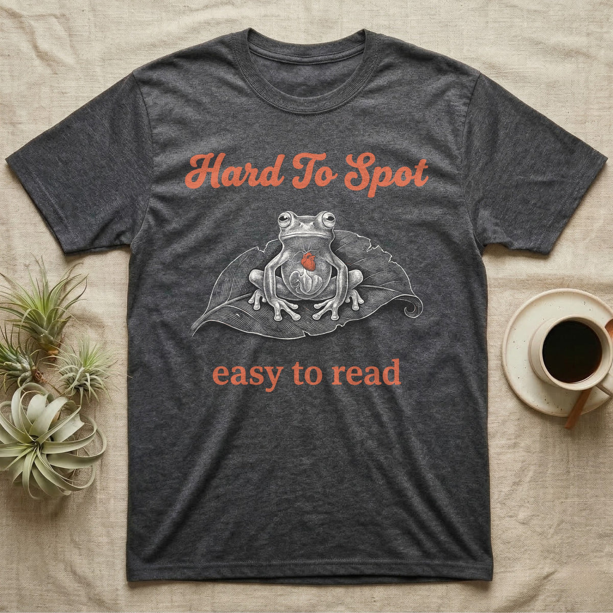 Glass Frog Engraving T-Shirt in Dark Heather — White-line engraving of a glass frog sitting on a leaf with a coral heart showing through its belly, between Hard To Spot and easy to read
