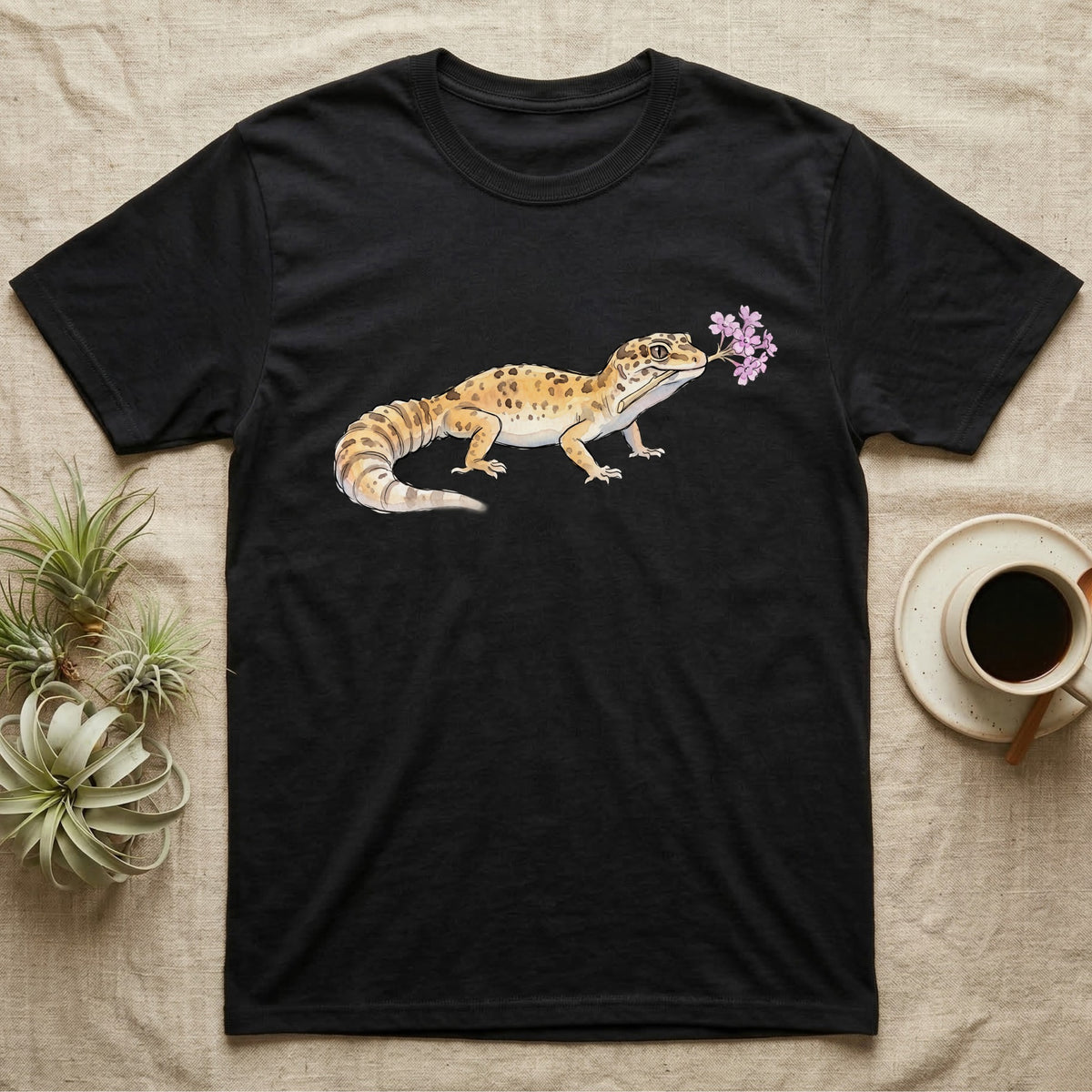 Leopard Gecko Blossom T-Shirt in Black — Watercolor illustration of a sandy-gold spotted leopard gecko holding a sand verbena blossom sprig, on a dark grey t-shirt.