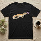 Leopard Gecko Blossom T-Shirt in Black — Watercolor illustration of a sandy-gold spotted leopard gecko holding a sand verbena blossom sprig, on a dark grey t-shirt.