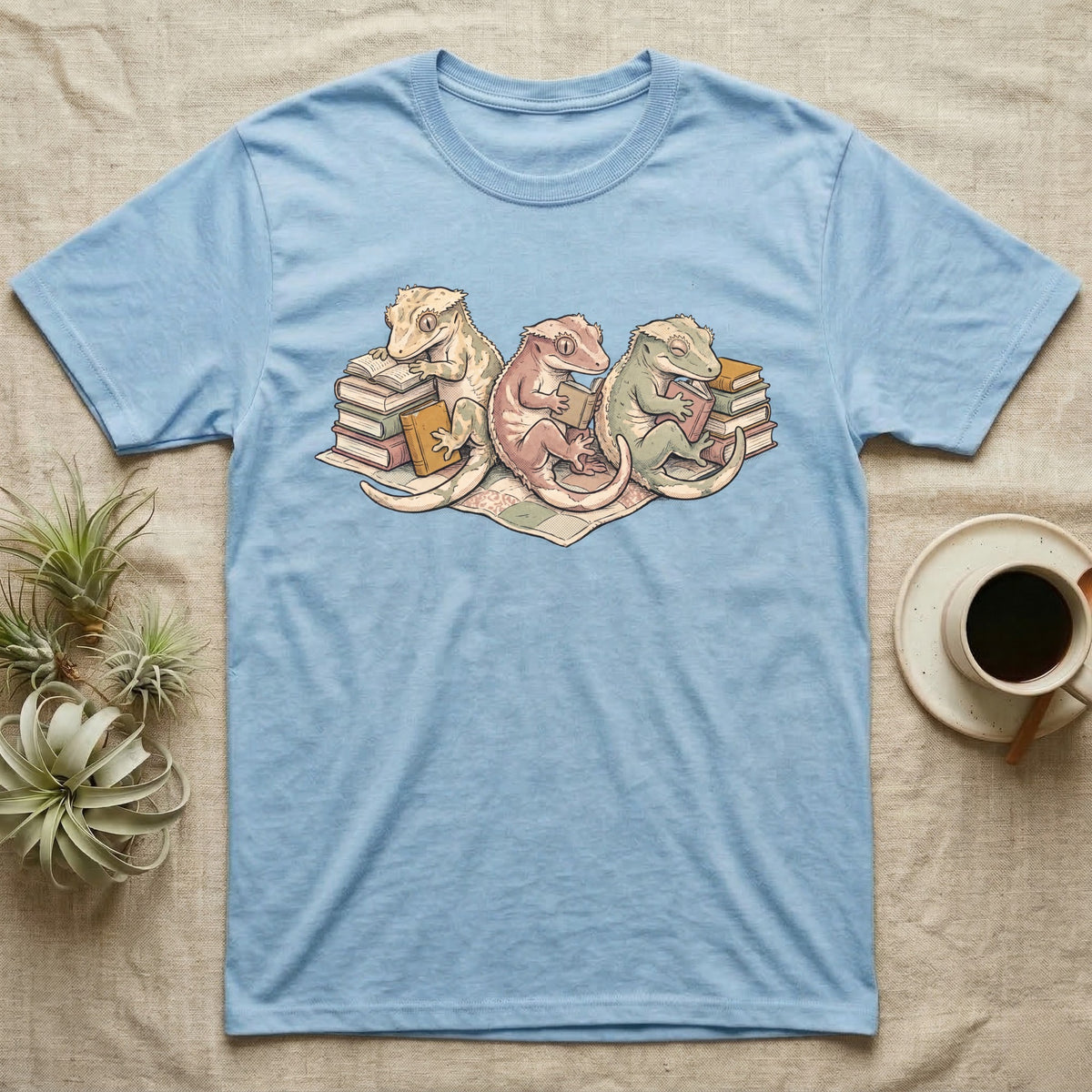 Gecko Book Club T-Shirt in Light Blue — Warm storybook illustration of three crested geckos in a cozy reading row, each beside a tiny stack of books.