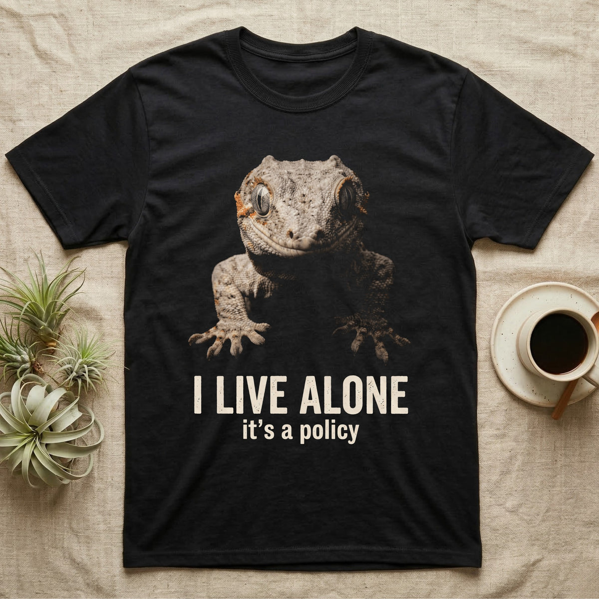Deadpan photographic mugshot of a gargoyle gecko reaching toward the camera above the distressed text I Live Alone, it's a policy. — shown on a Black tee.