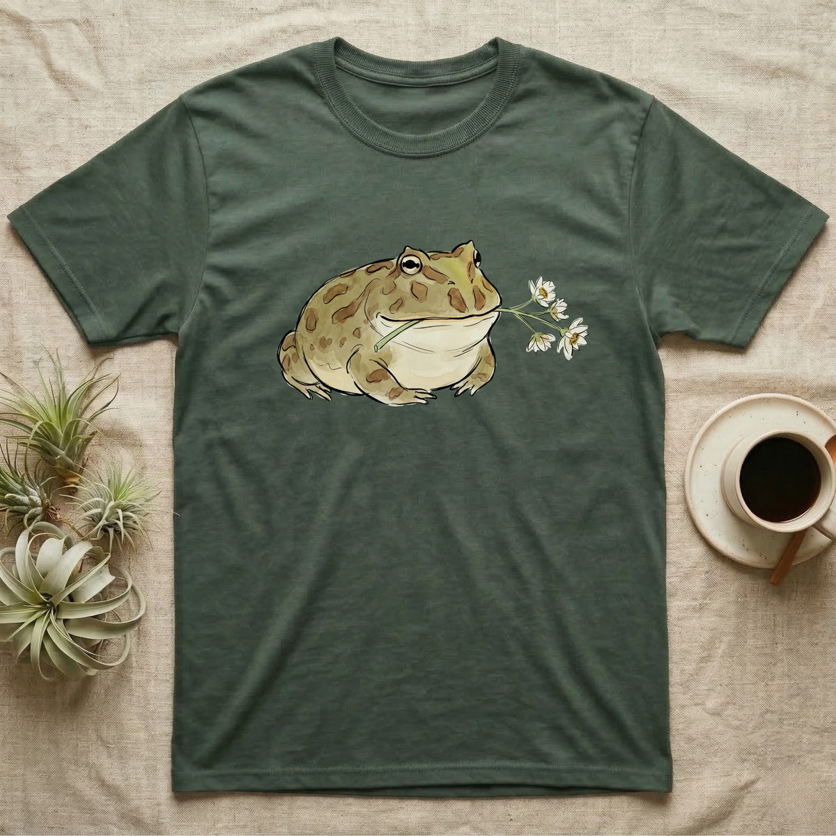 Pacman Frog Blossom T-Shirt in Military Green — Watercolor illustration of an olive-tan Pacman frog holding a water-lily blossom sprig, on a dark grey t-shirt.