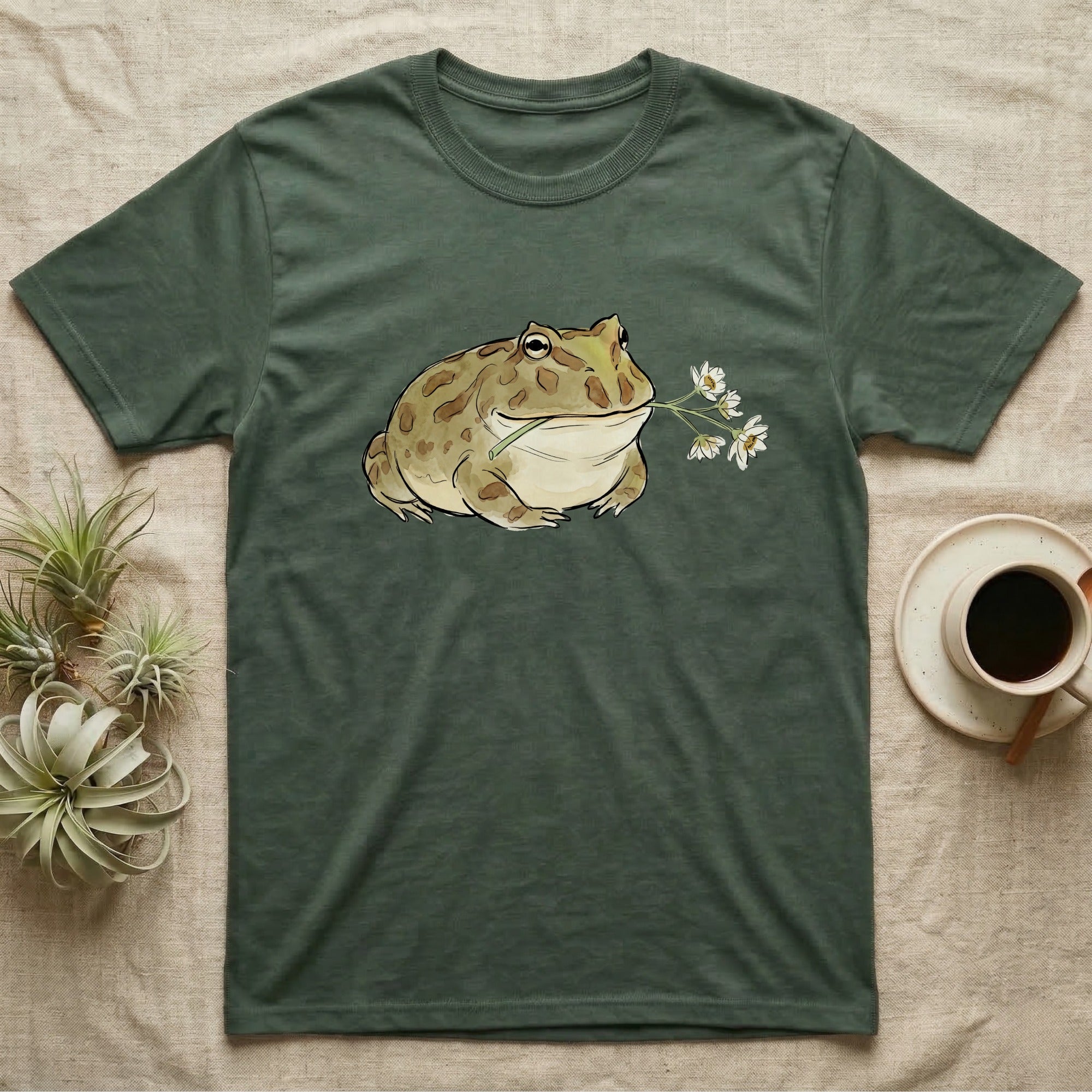 Pacman Frog Blossom T-Shirt in Military Green — Watercolor illustration of an olive-tan Pacman frog holding a water-lily blossom sprig, on a dark grey t-shirt.