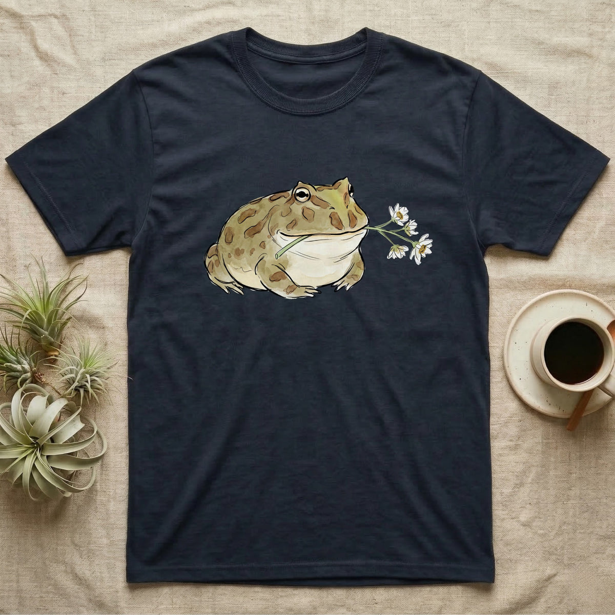 Pacman Frog Blossom T-Shirt in Navy — Watercolor illustration of an olive-tan Pacman frog holding a water-lily blossom sprig, on a dark grey t-shirt.