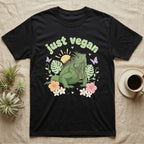 Green Iguana Baby T-Shirt in Black — Green iguana with spiky crest and dewlap among pastel leaves and flowers under puffy green bubble text "just vegan".