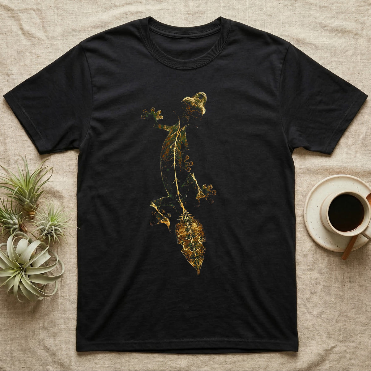 Full-body illustration of an eyelash leaf-tailed gecko mid-climb with a mottled olive-and-gold leaf-shaped tail blending into camouflage texture, no on-shirt text. — shown on a Black tee.