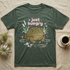 African Bullfrog Baby T-Shirt in Military Green — Pastel African bullfrog eyeing a tiny bug among reeds and flowers, puffy pink bubble wordmark "just hungry" above.